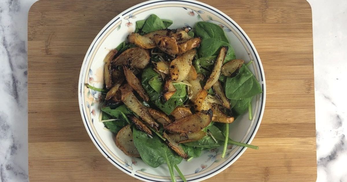 A close-up photo of a round, white bowl filled with spinach and grilled pear with balsamic reduction on a light brown wooden cutting board.