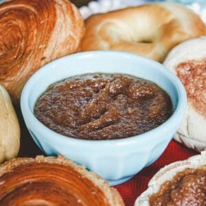 Closeup of a white bowl holding creamy, dark apple butter, surrounded by different breakfast breads.