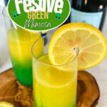 A glass of festive green mimosa with a lemon slice on the rim sits on a white counter.