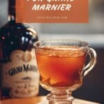A glass cup filled with Grand Marnier, next to a bottle with a black and gold label that says "Grand Marnier" on a wooden table.