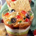 Clear individual cups filled with layers of seven-layer dip, topped with cheese, olives, and tomatoes.