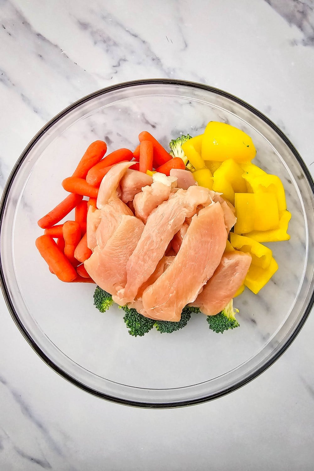 Raw sliced chicken breast added to a glass bowl with baby carrots, broccoli, and yellow bell pepper