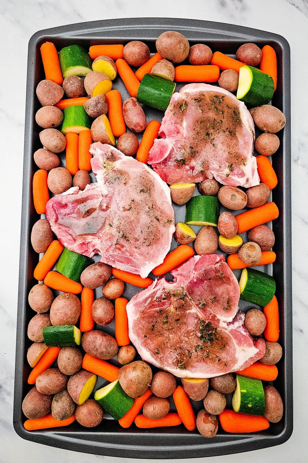 Fully assembled sheet pan with glazed pork chops surrounded by baby carrots, red potatoes, and zucchini ready to bake