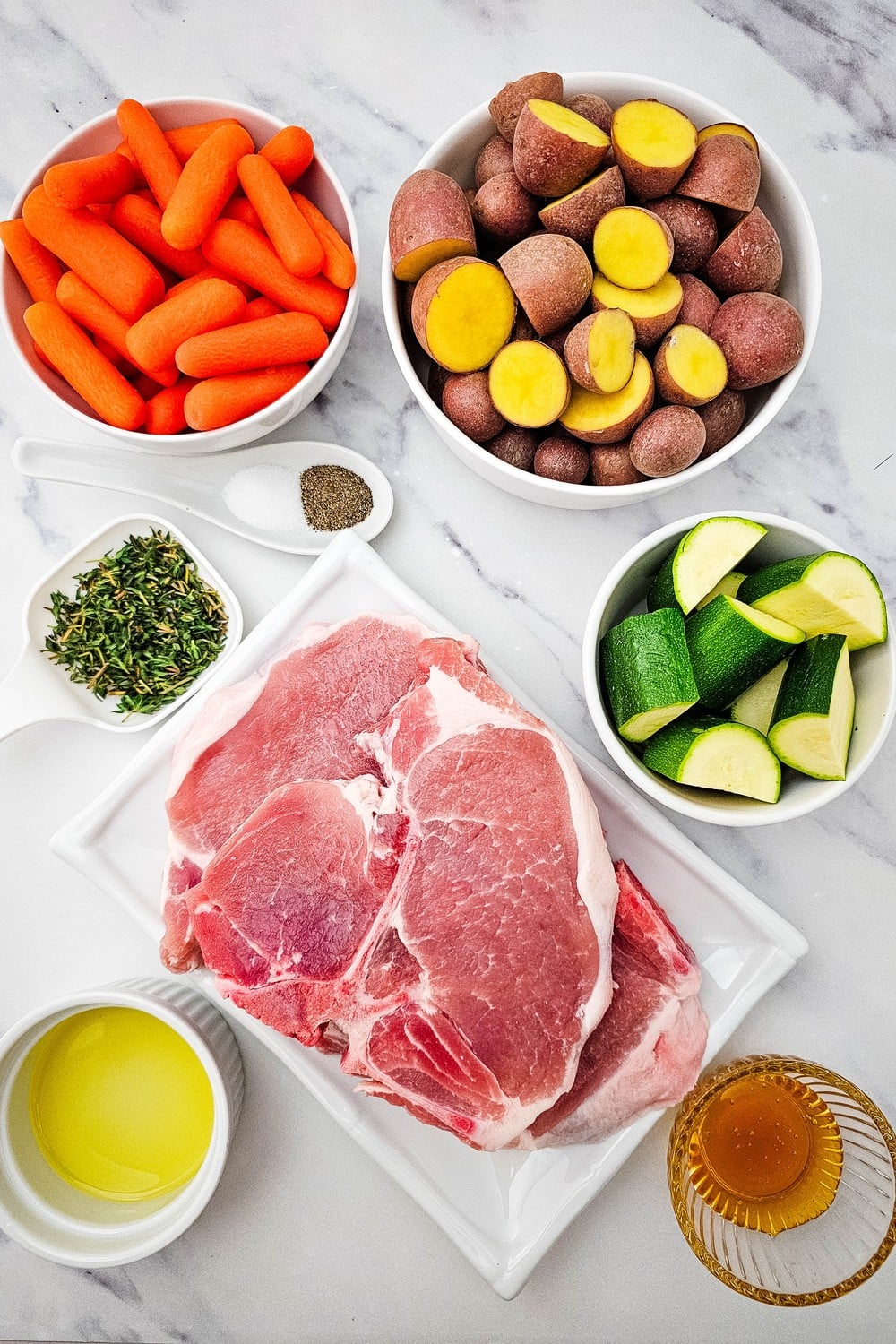 Ingredients flat lay: raw bone-in pork chops, baby carrots, halved red potatoes, zucchini chunks, olive oil, honey, and thyme