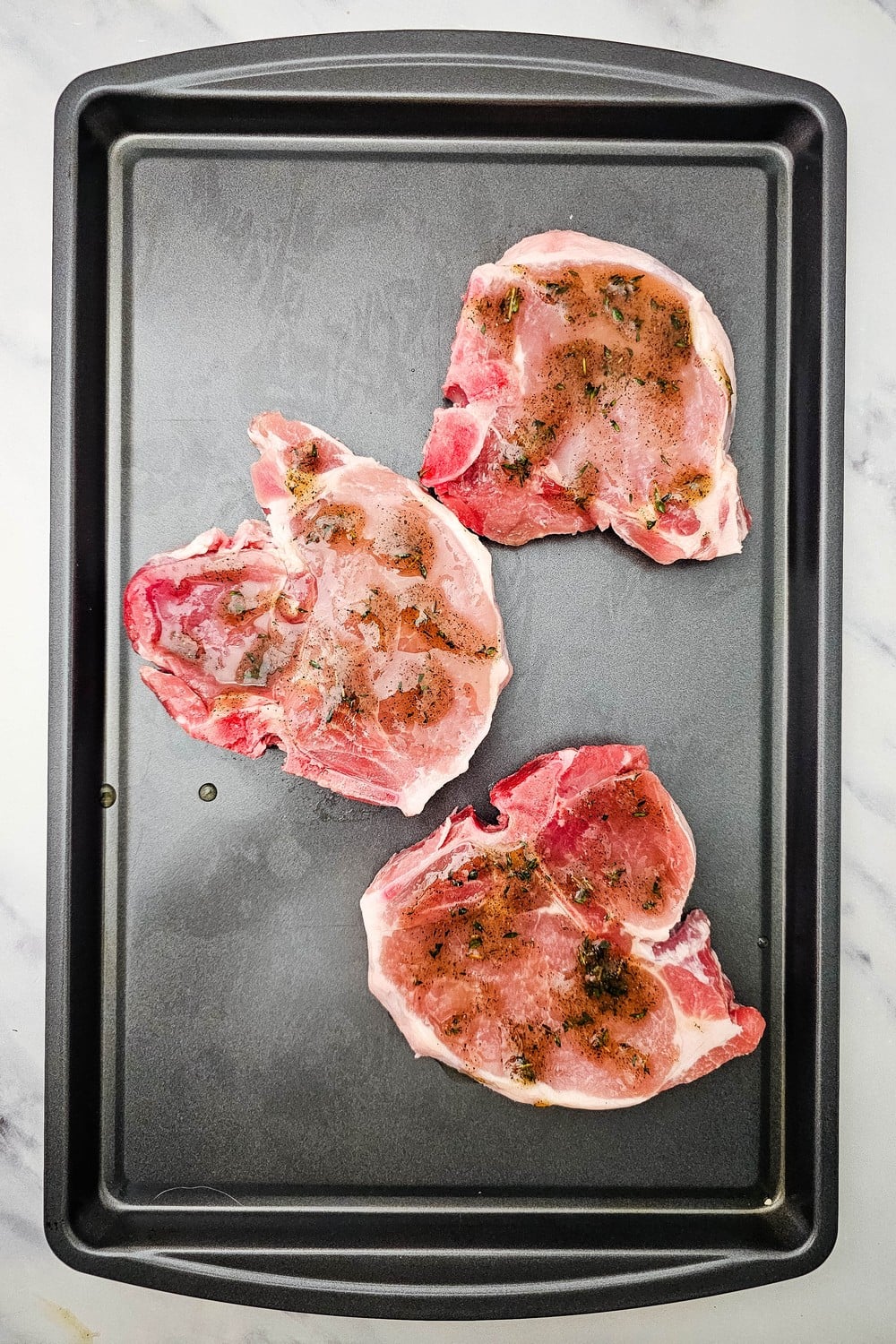 Three raw bone-in pork chops on sheet pan brushed with honey thyme glaze