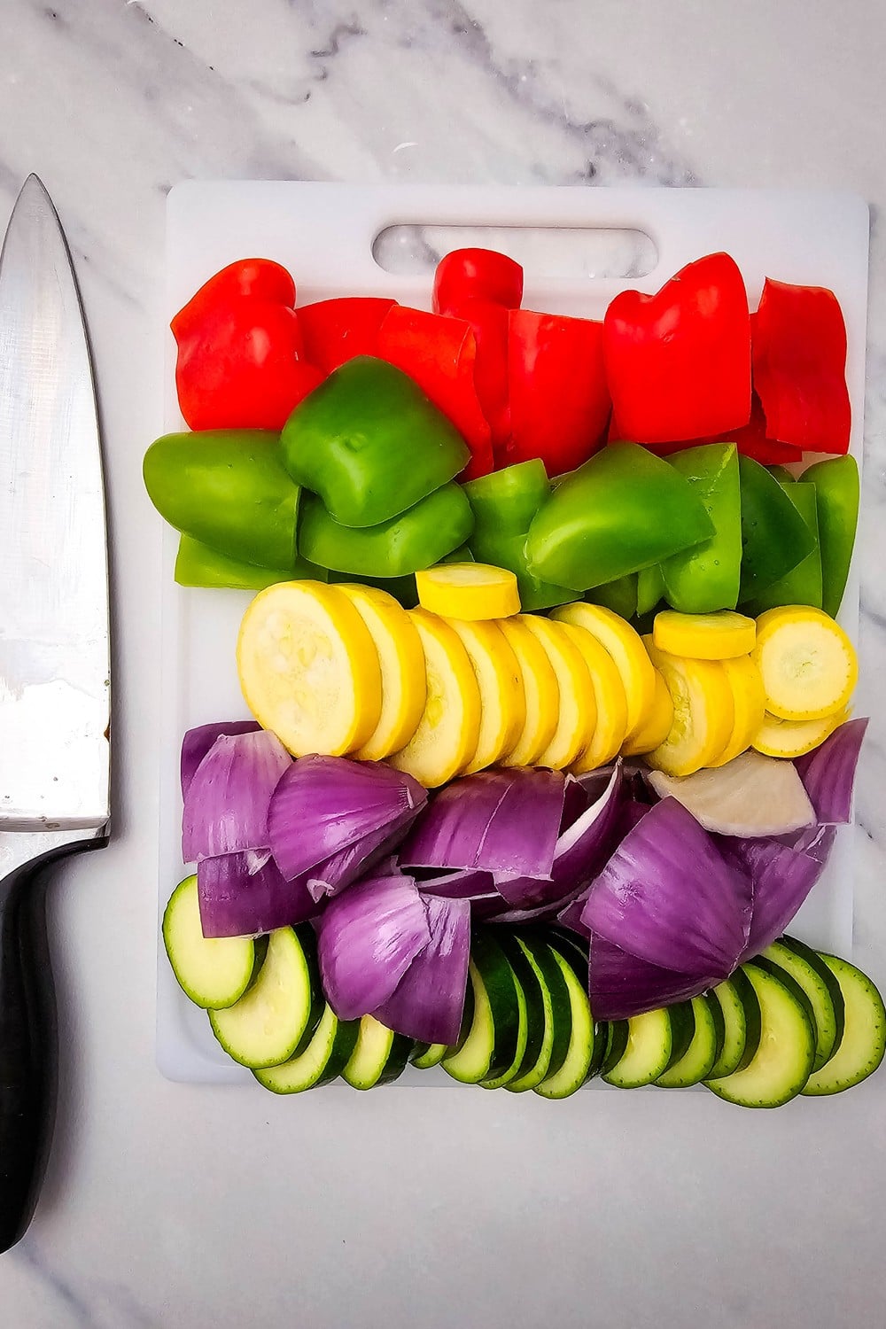 Chopped red and green bell peppers, yellow squash, red onion, and zucchini on cutting board