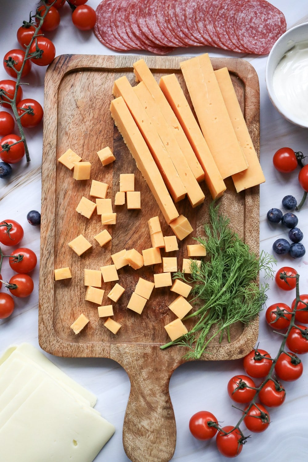 Prepping cheddar cheese cubes and sticks on a wooden cutting board for a 4th of July charcuterie board with surrounding tomatoes, blueberries, salami, and dill
