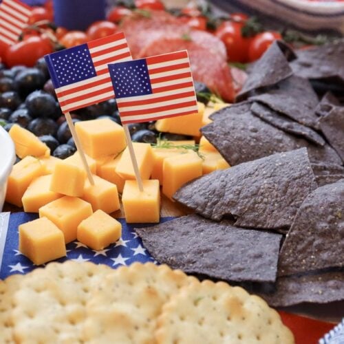 4th of July charcuterie board close-up with American flag picks, cheddar cubes, blue corn chips, ranch dip, and crackers