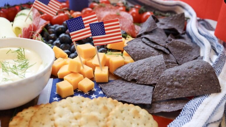 4th of July charcuterie board close-up with American flag picks, cheddar cubes, blue corn chips, ranch dip, and crackers
