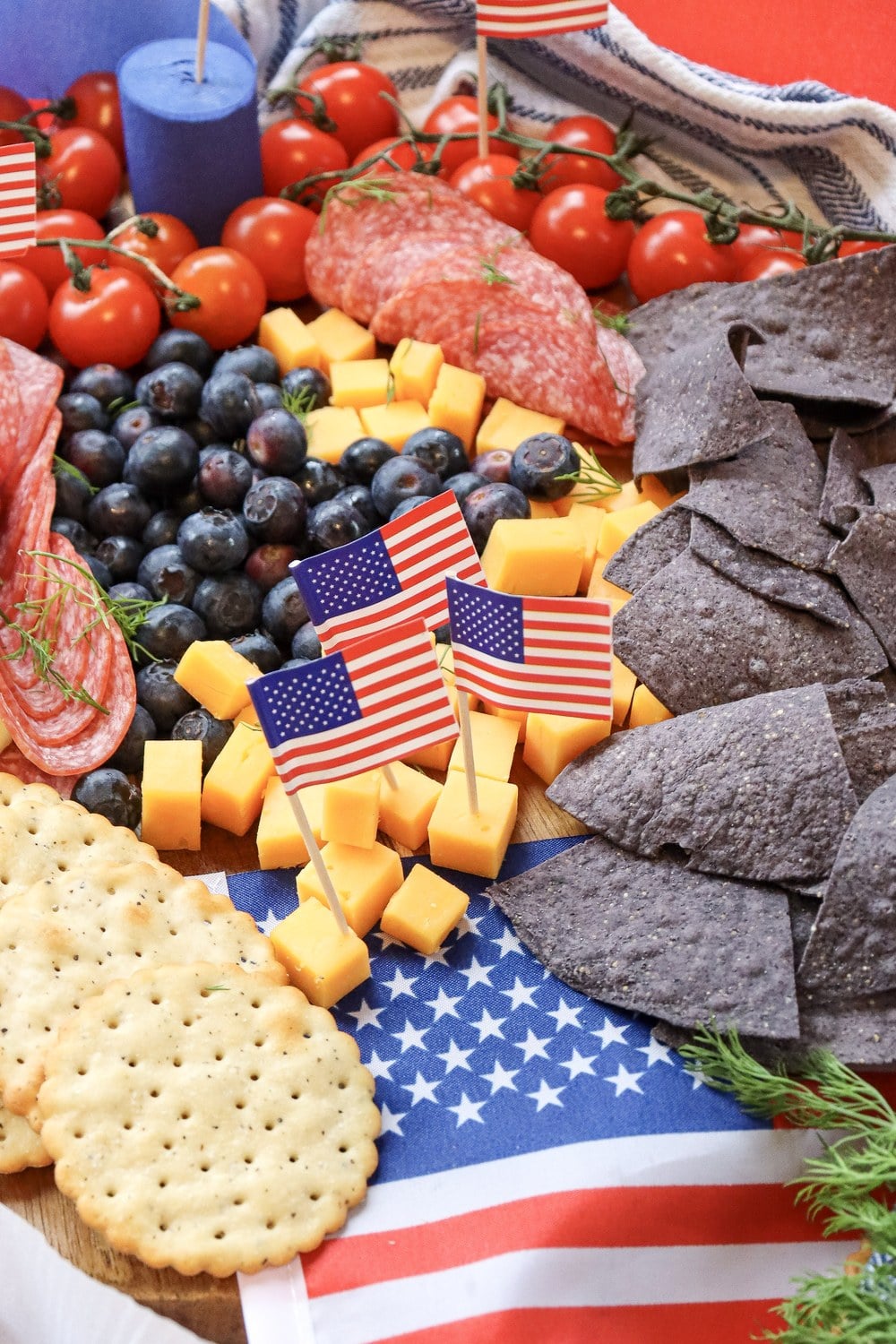 Side angle of a 4th of July charcuterie board with cherry tomatoes, swiss cheese, salami, blueberries, cheddar, blue corn chips, and crackers
