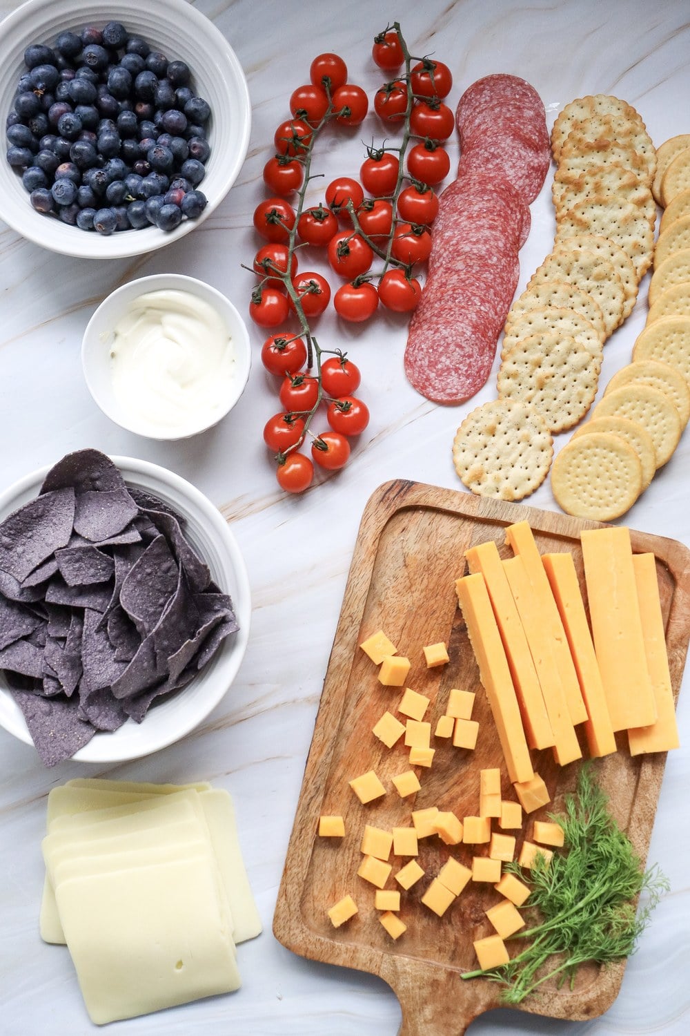 4th of July charcuterie board ingredients flat lay with cherry tomatoes, blueberries, cheddar, swiss, salami, crackers, blue corn chips, and ranch dip