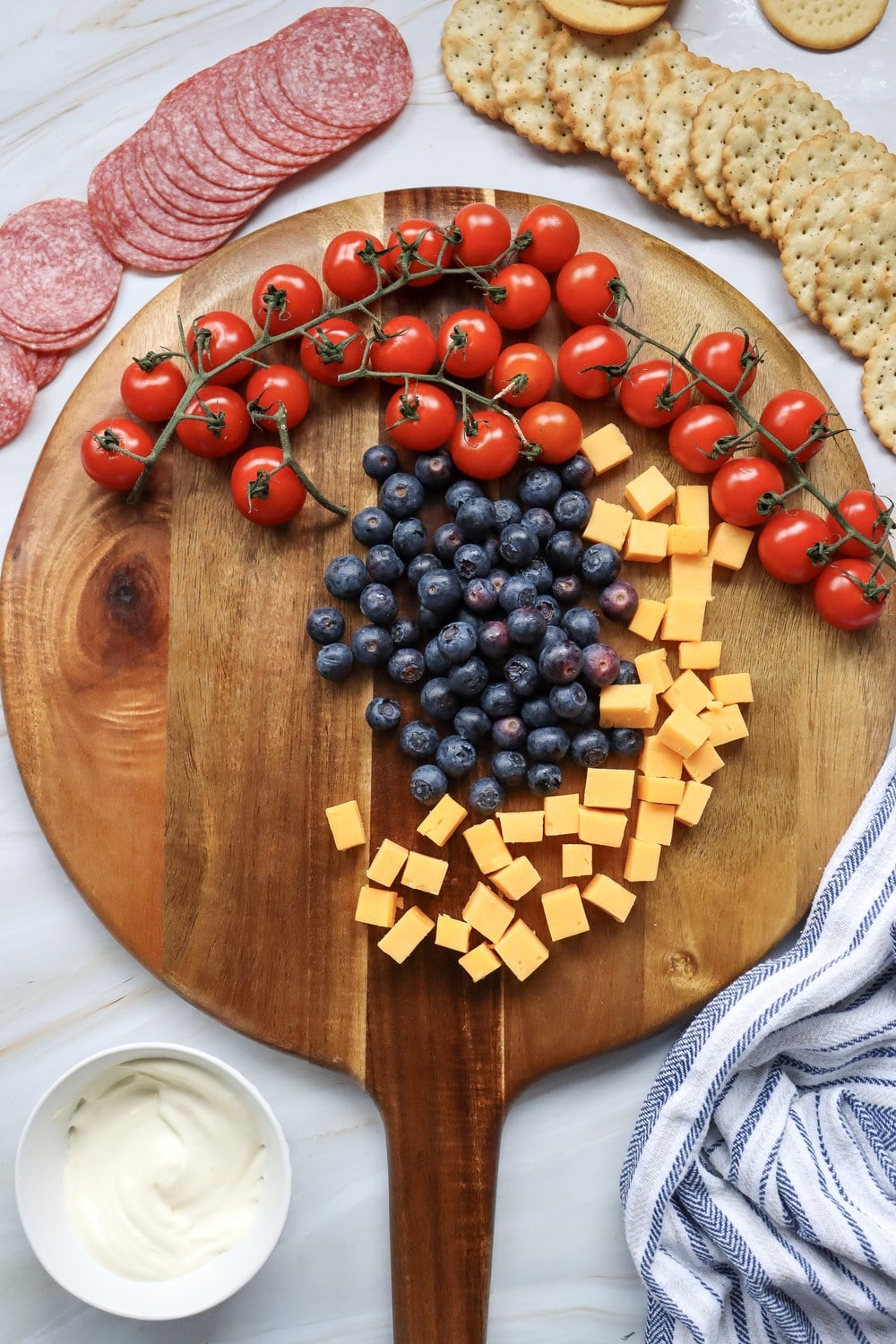 Starting to assemble a 4th of July charcuterie board with cherry tomatoes on the vine, blueberries, cheddar cubes, and salami on a round wooden board