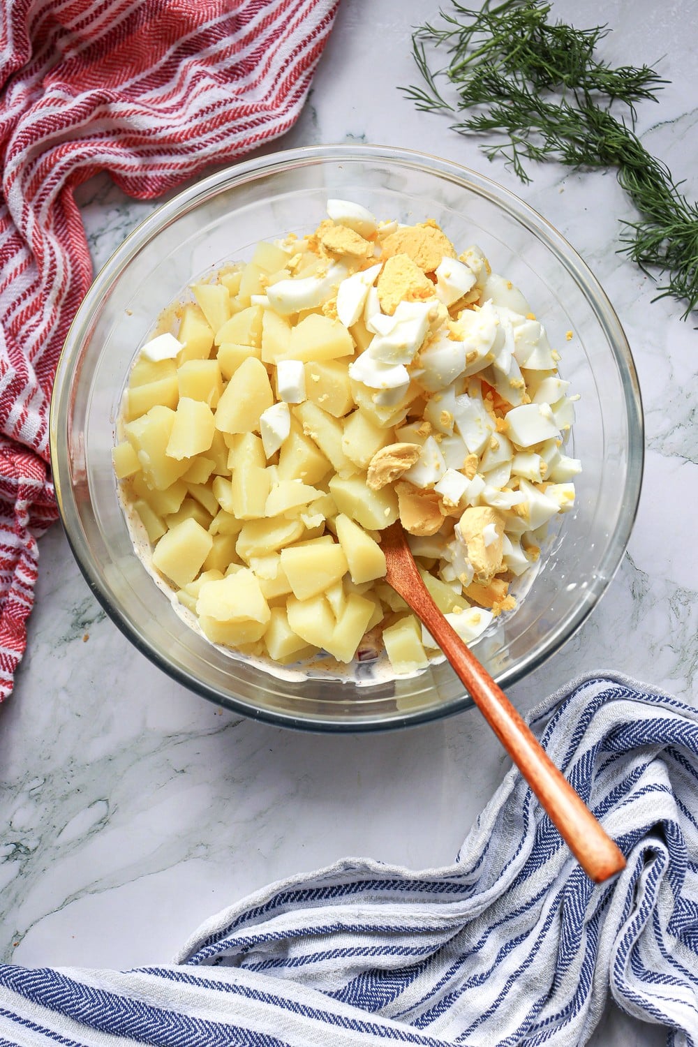 Folded 4th of July potato salad with potatoes, dressing, and chopped hard-boiled eggs in a bowl with a wooden spoon