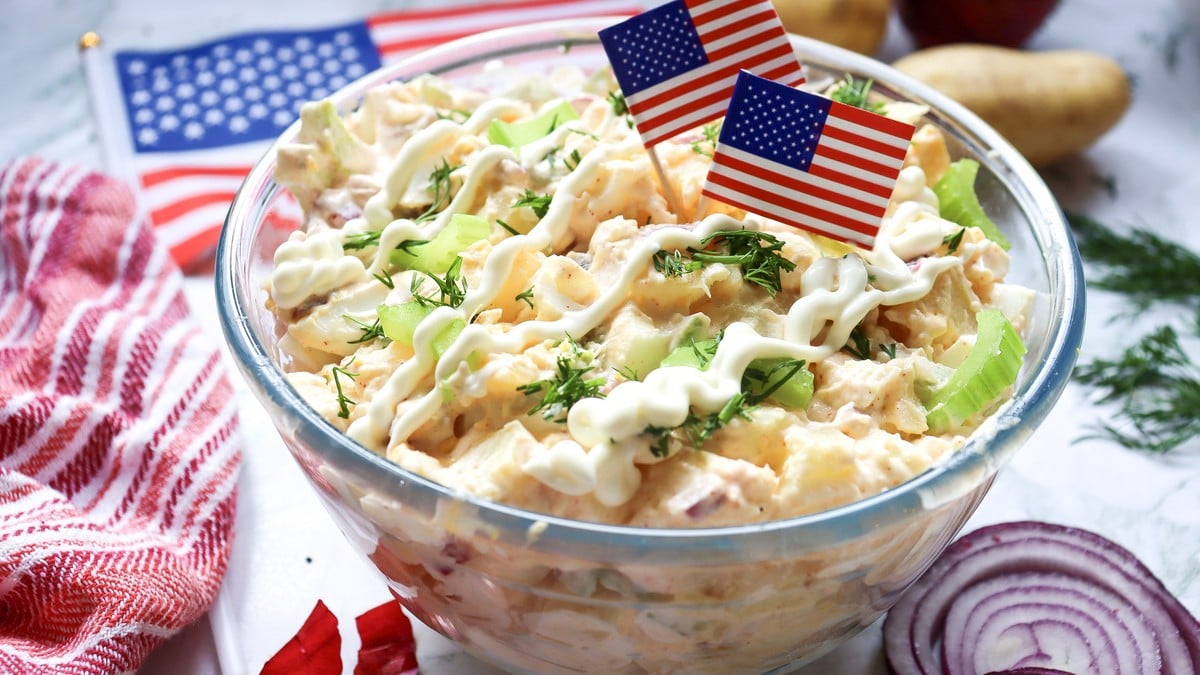 Wide overhead shot of 4th of July potato salad in a white bowl surrounded by small American flags and fresh dill