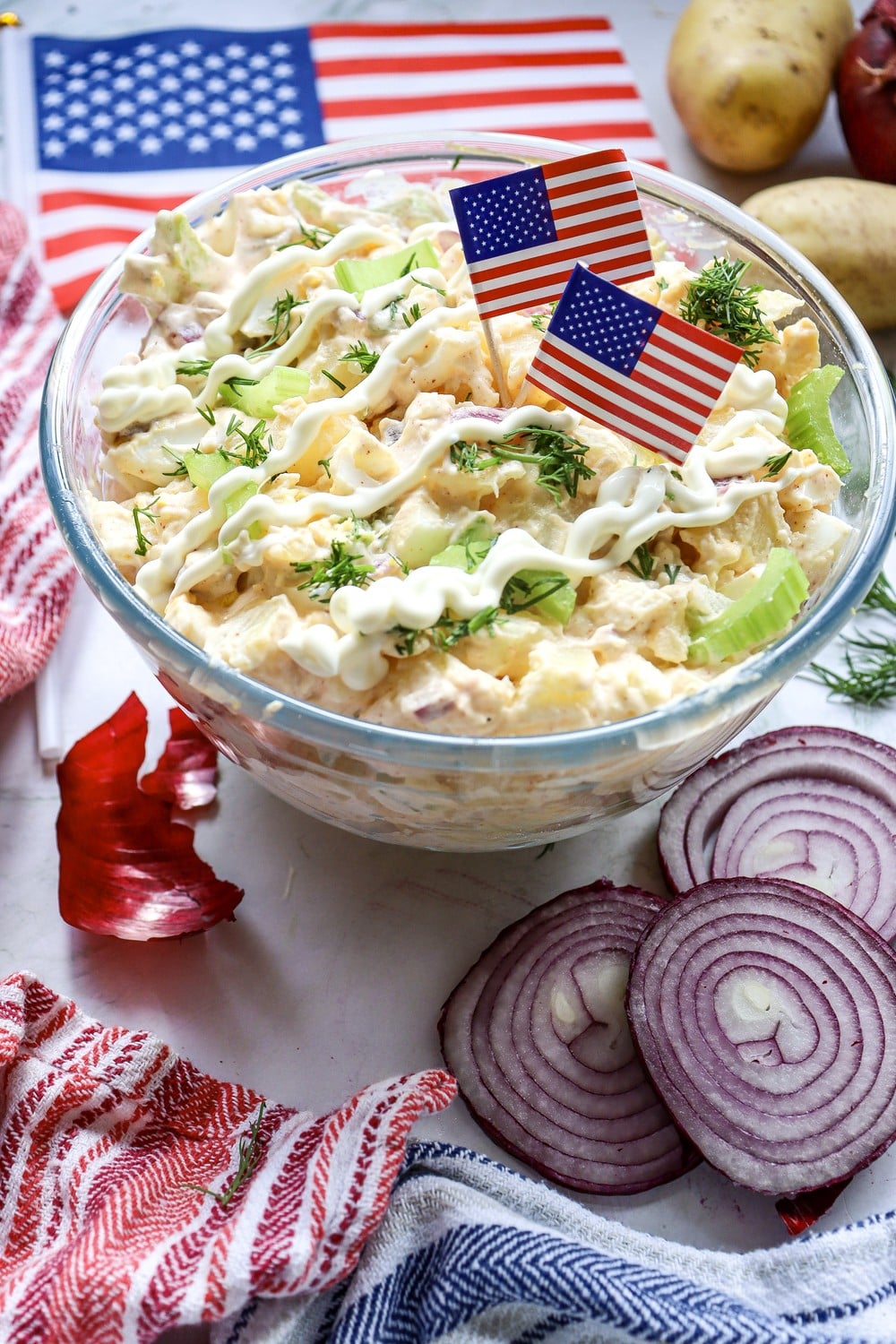 Overhead view of 4th of July potato salad in a white bowl with small American flags, sliced red onion, and fresh dill garnish