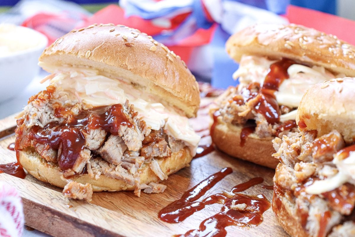 Three BBQ pulled pork sandwiches with coleslaw on toasted buns staged on a wooden board with a red, white, and blue 4th of July background.