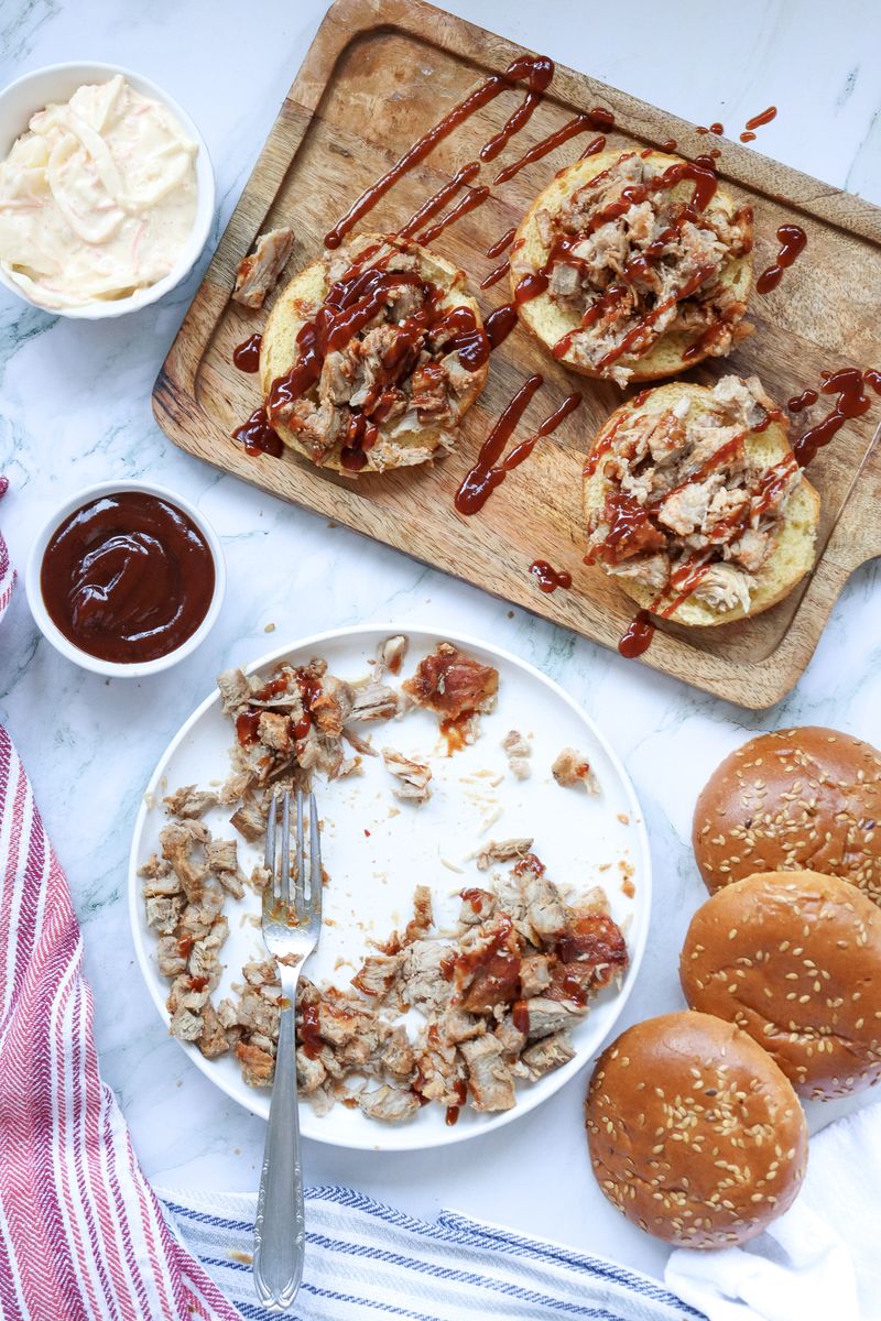 Open-faced BBQ pulled pork sandwiches on a wooden board with BBQ sauce drizzled on top, ready for the coleslaw to be added.