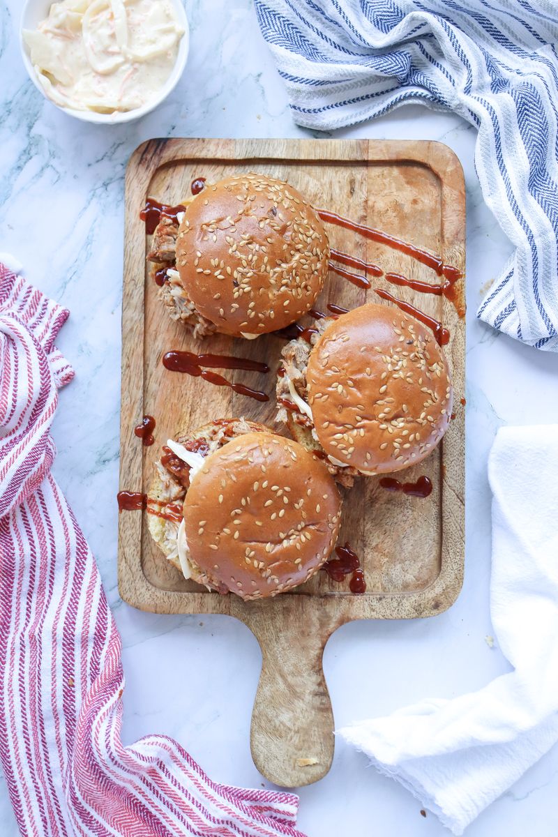 Three BBQ pulled pork sandwiches with coleslaw arranged on a rustic wooden board, ready to serve at a backyard cookout.