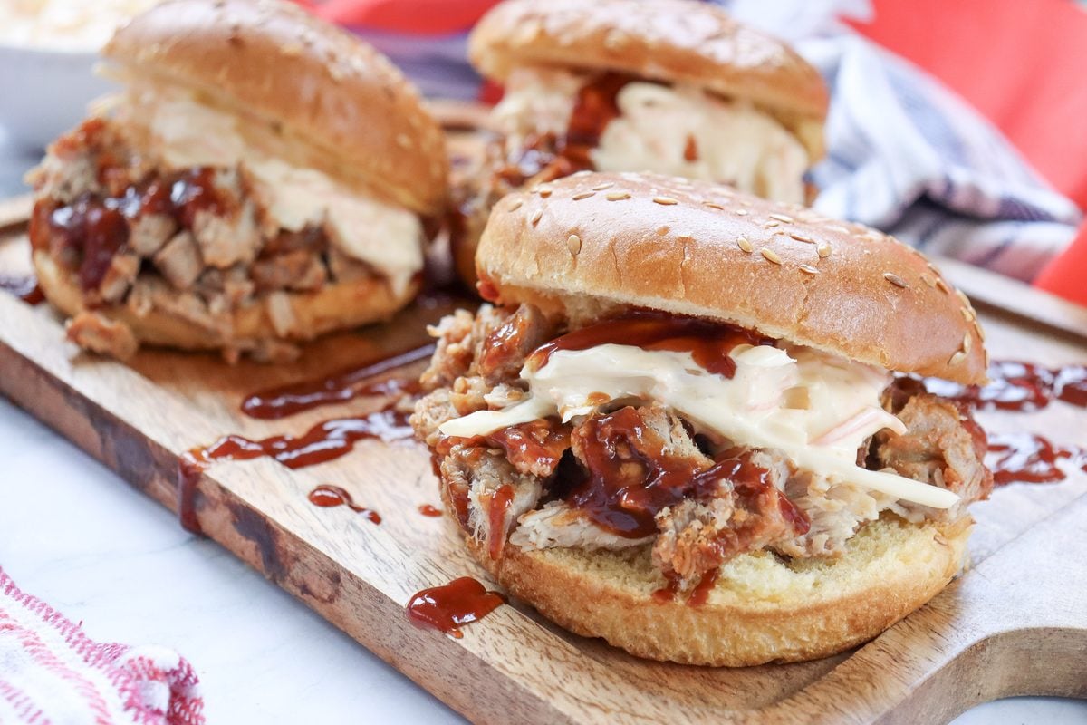 BBQ pulled pork sandwiches with coleslaw and dripping BBQ sauce styled on a wooden board with red, white, and blue 4th of July decor.