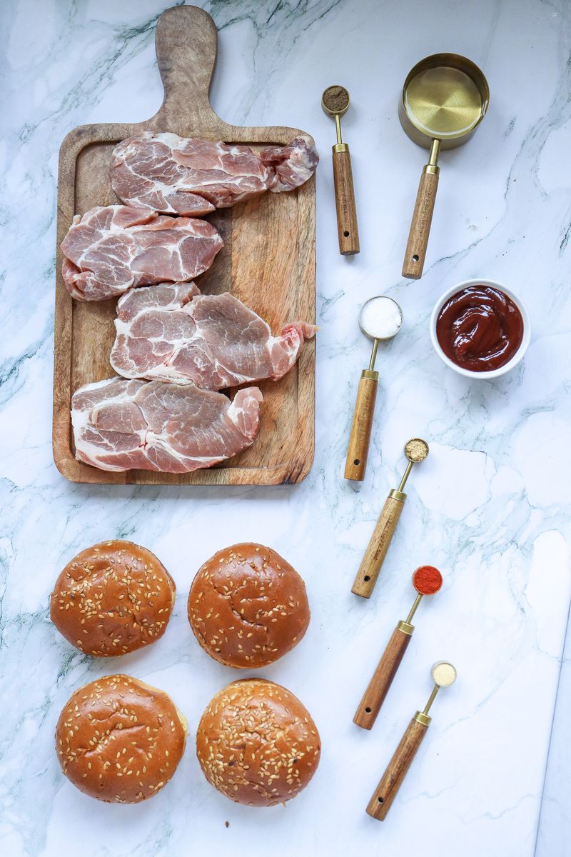 Flat lay of raw pork shoulder on a wooden board with sesame buns, BBQ sauce in a small bowl, and measuring cups of spices for slow cooker BBQ pulled pork sandwiches.