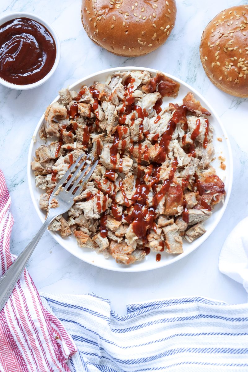Plate of shredded BBQ pulled pork drizzled with extra BBQ sauce, ready to be piled onto toasted sandwich buns.