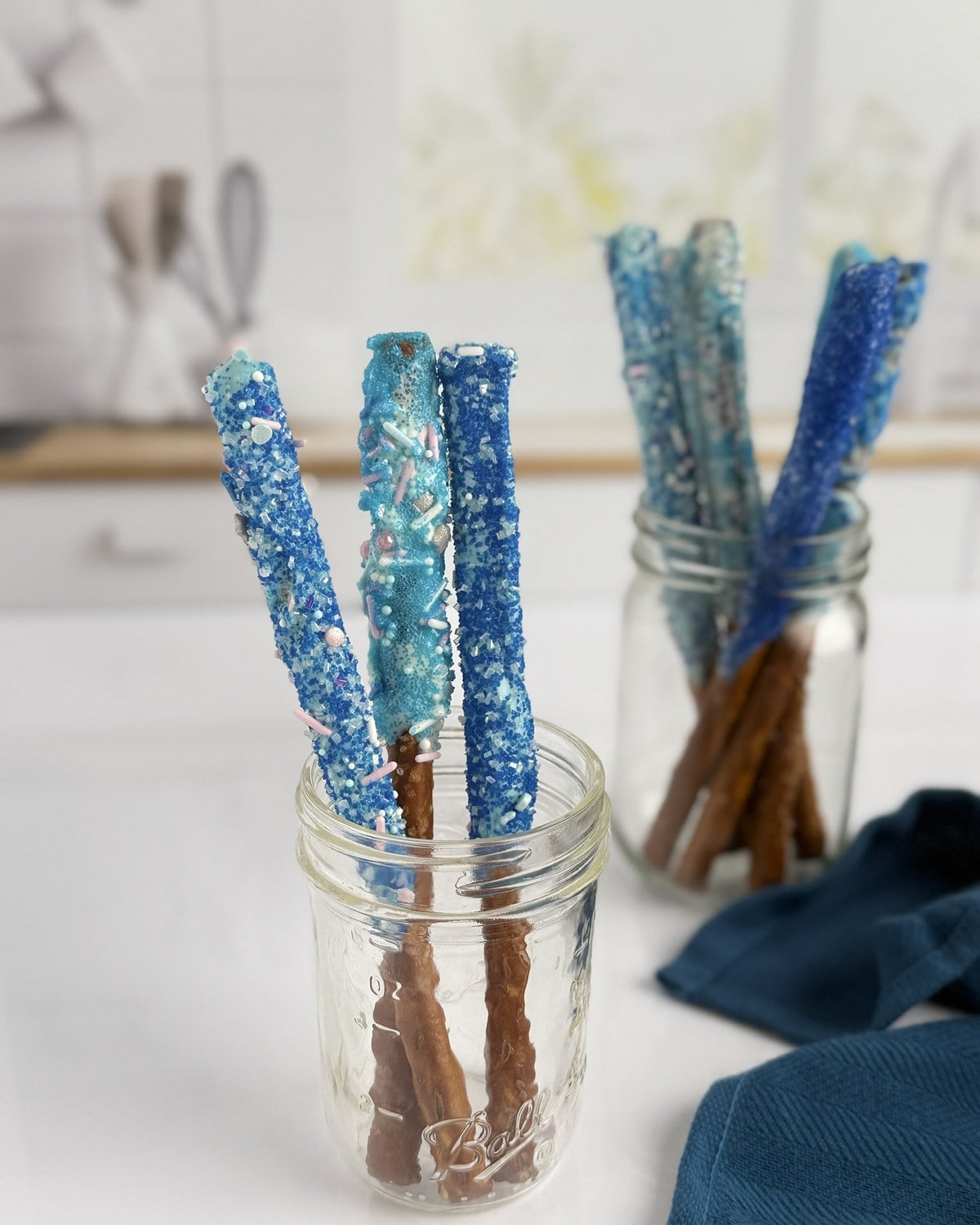 Two clear mason jars holding blue-dipped pretzel rods against a soft white bokeh background