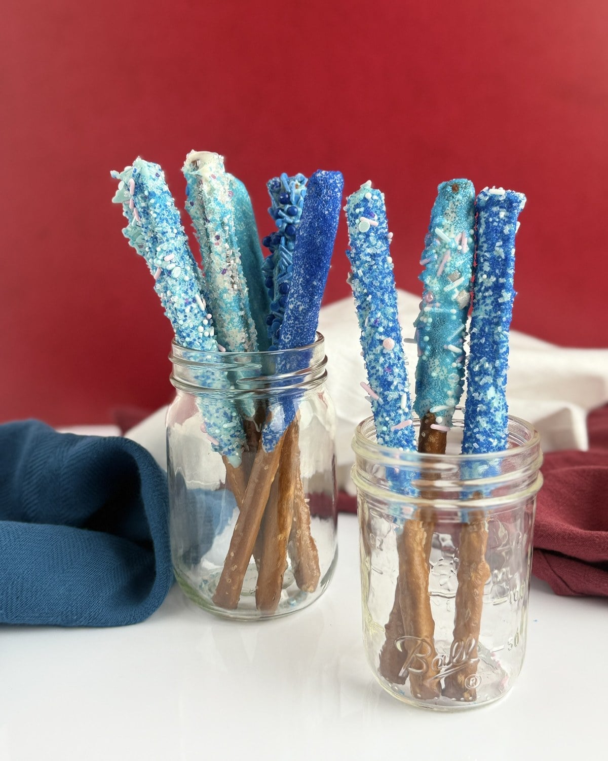 Two glass mason jars filled with blue-dipped pretzel rods on a red backdrop with navy and burgundy cloth napkins