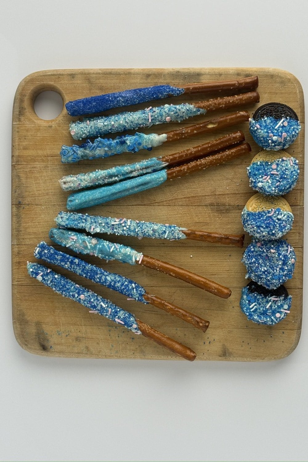 Overhead board of blue-dipped pretzel rods and blue-dipped sandwich cookies on a wooden cutting board