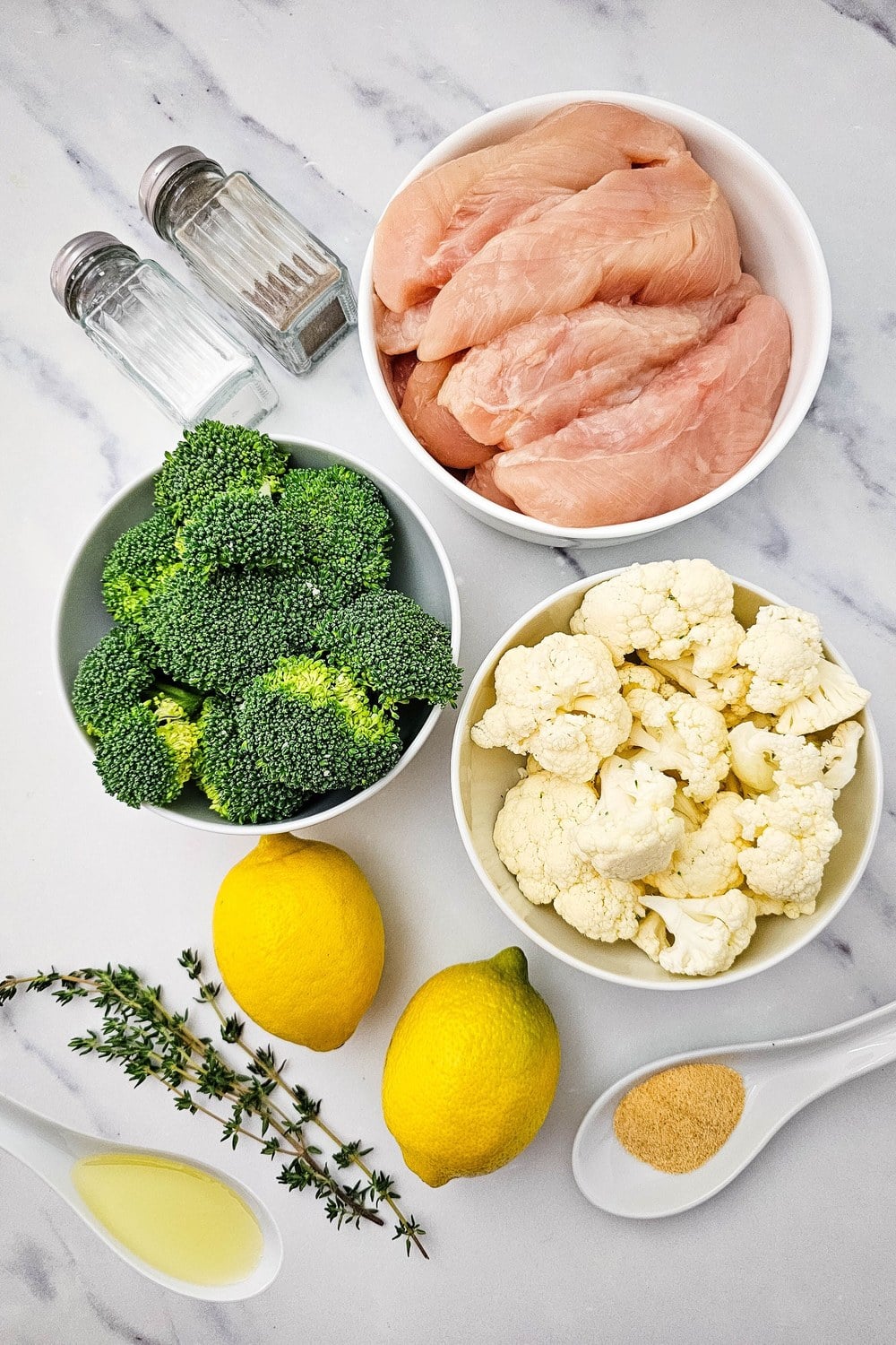Overhead flat lay of ingredients for sheet pan lemon chicken and veggies including chicken tenderloins, broccoli, cauliflower, lemons, fresh thyme, olive oil, garlic powder, salt, and pepper.