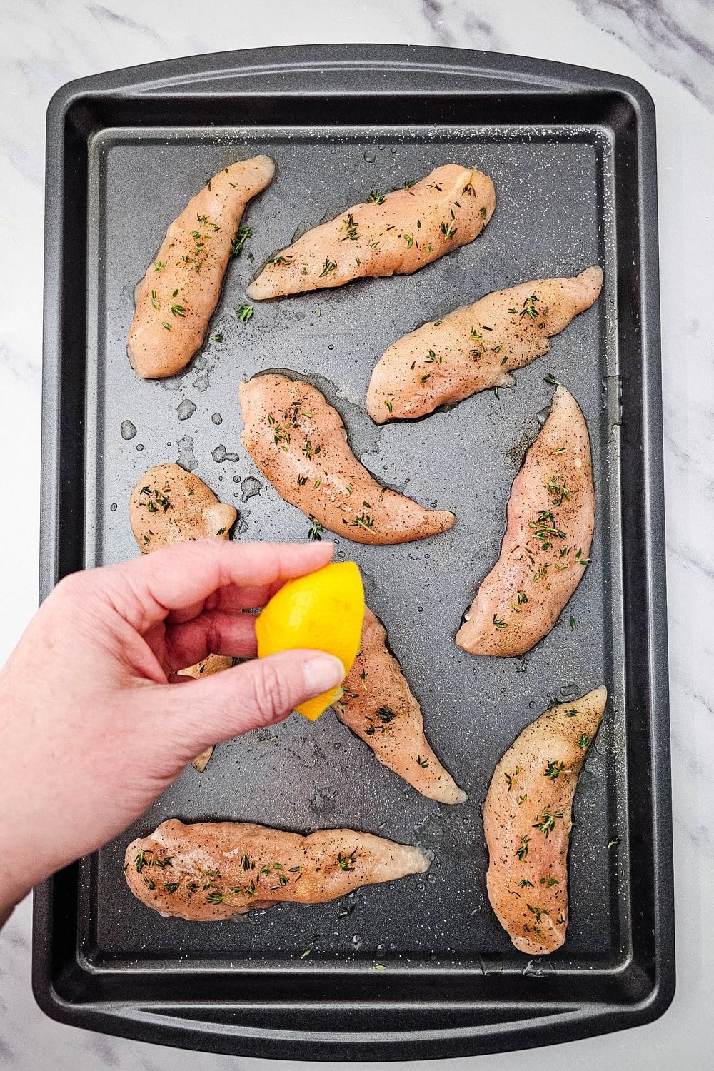 A hand squeezing half a lemon over seasoned chicken tenderloins on a sheet pan.