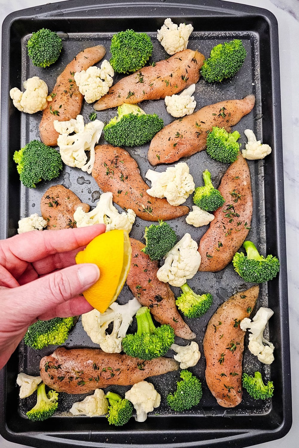 A hand squeezing half a lemon over the assembled chicken and vegetables on a sheet pan.