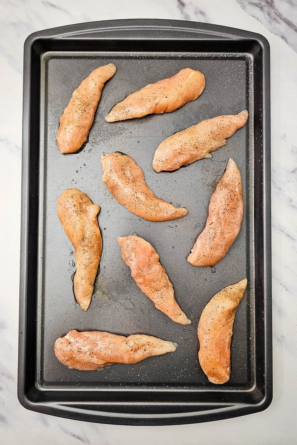 Raw chicken tenderloins arranged on a sheet pan and lightly seasoned with salt and pepper.
