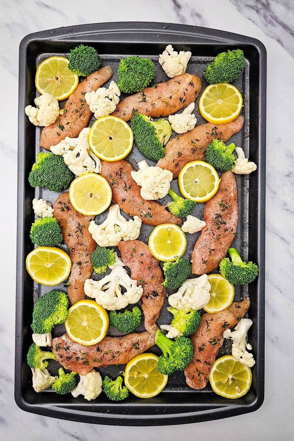 Sheet pan with chicken tenderloins, broccoli, cauliflower, and thin lemon slices ready to bake.