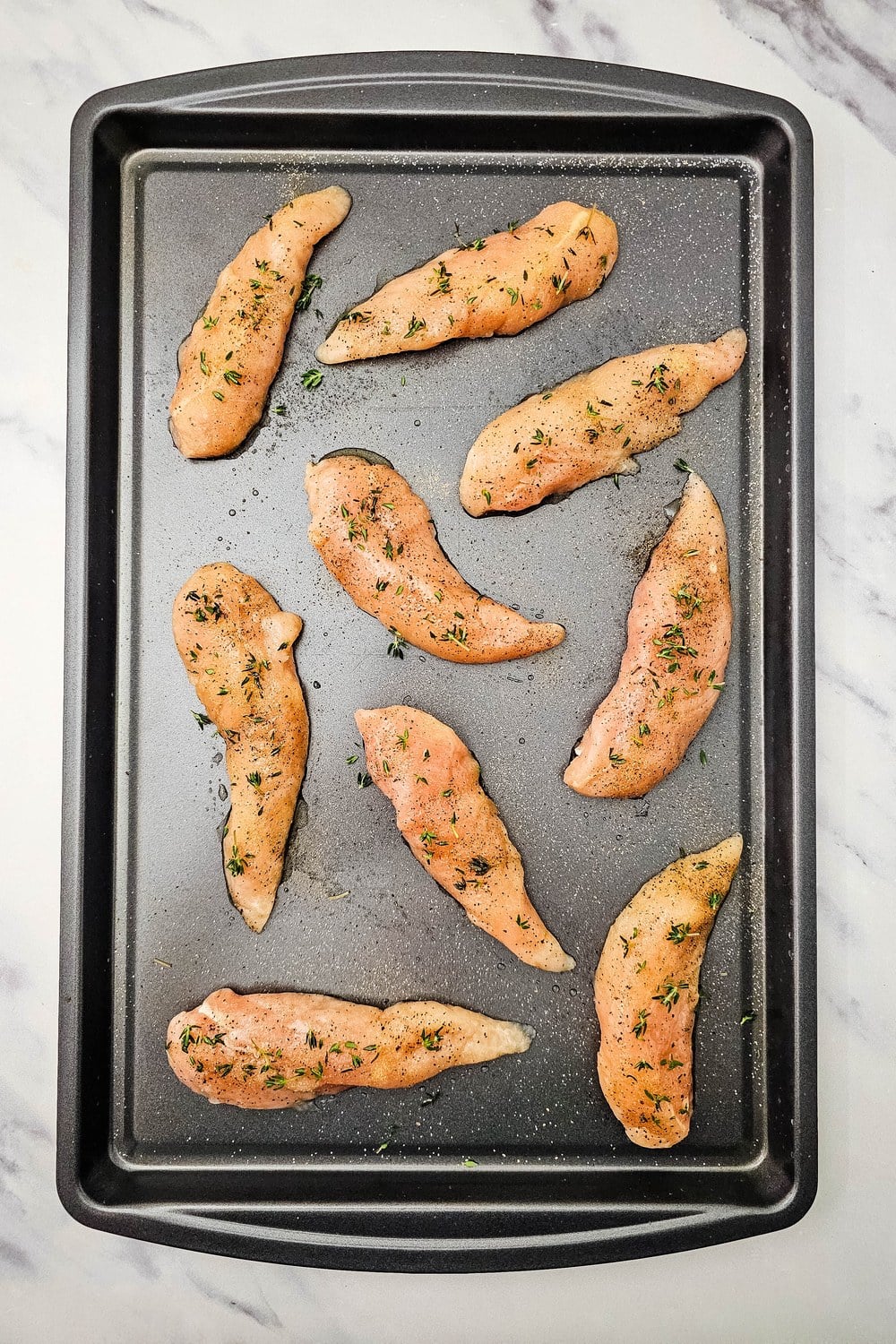 Seasoned chicken tenderloins on a sheet pan sprinkled with fresh thyme leaves.