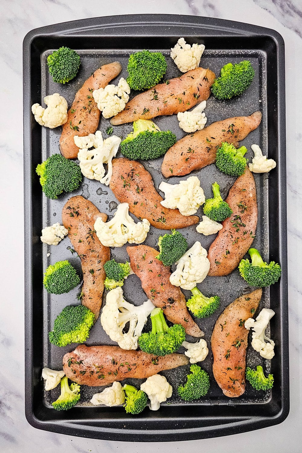 Sheet pan with seasoned chicken tenderloins surrounded by raw broccoli and cauliflower florets.