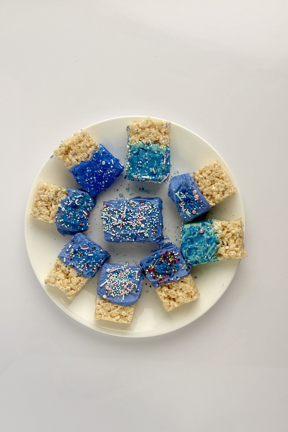 Overhead view of patriotic rice krispie treats arranged in a circle on a white plate, blue-dipped tops with mixed red white and blue sprinkles