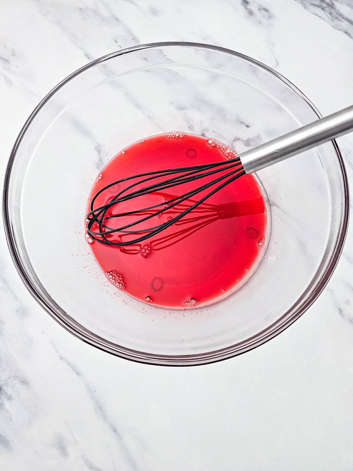 Strawberry Jell-O being whisked in a glass bowl until the gelatin powder dissolves