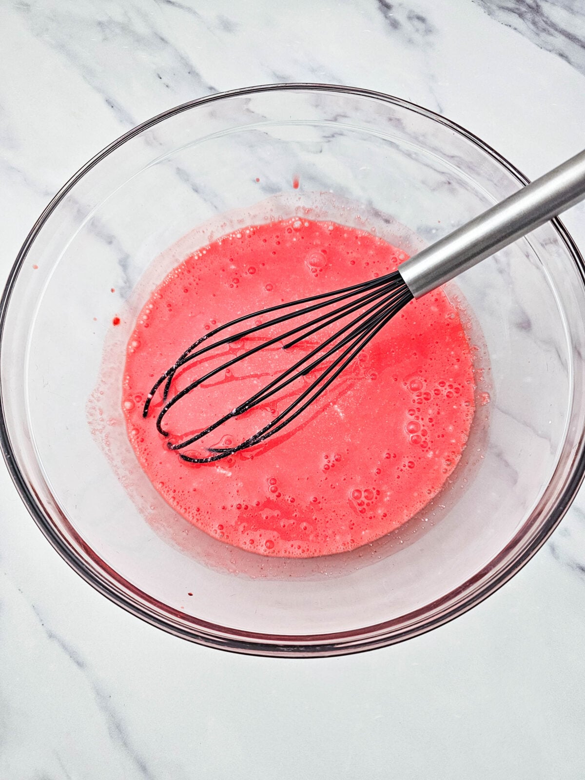 Fully dissolved strawberry gelatin sitting in a glass bowl with a whisk, ready to cool