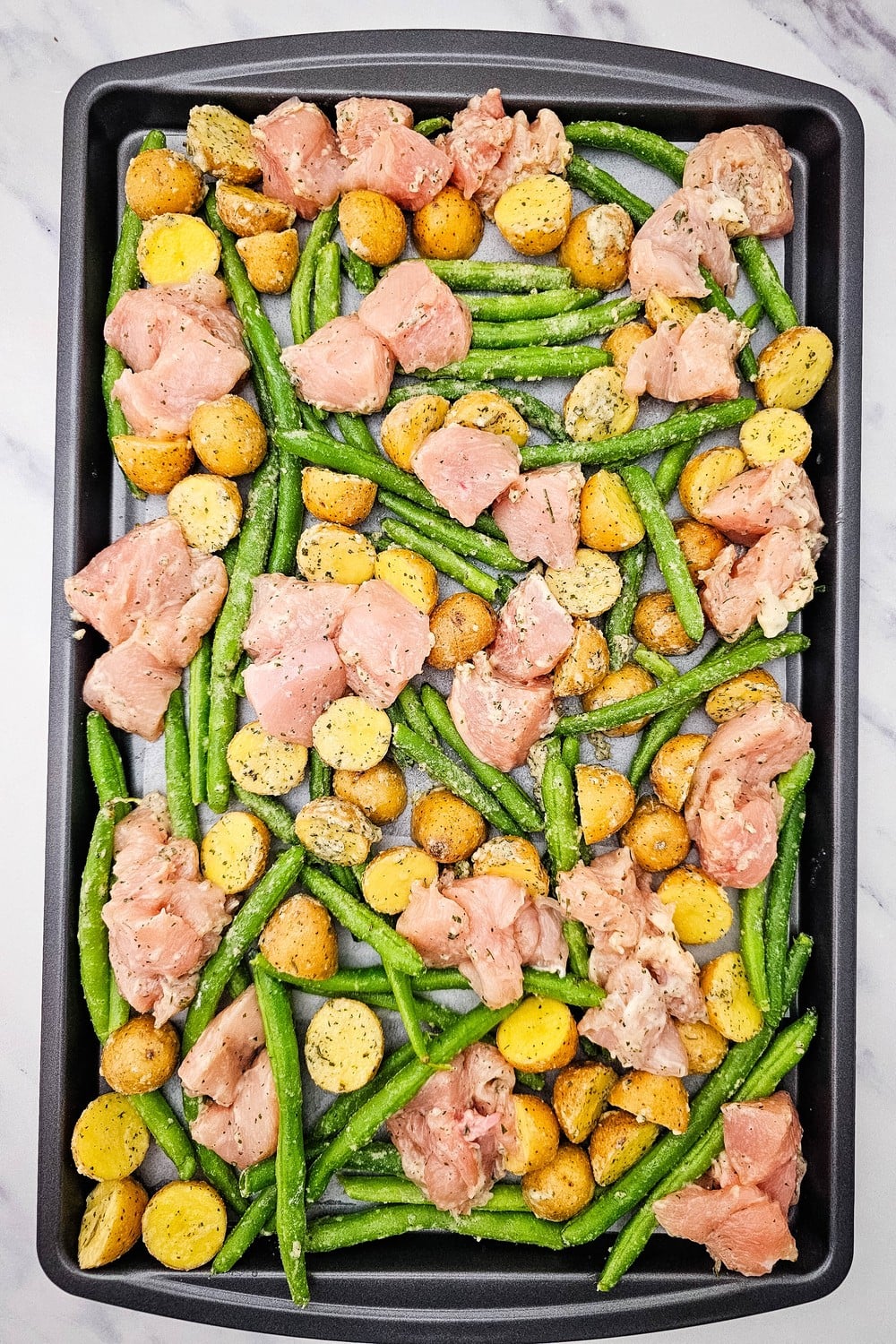 Sheet pan with seasoned chicken cubes arranged in open spaces between potatoes and green beans, ready for the oven.