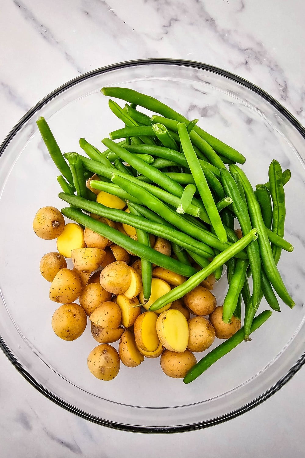 Halved baby potatoes and fresh green beans in a glass mixing bowl before tossing.