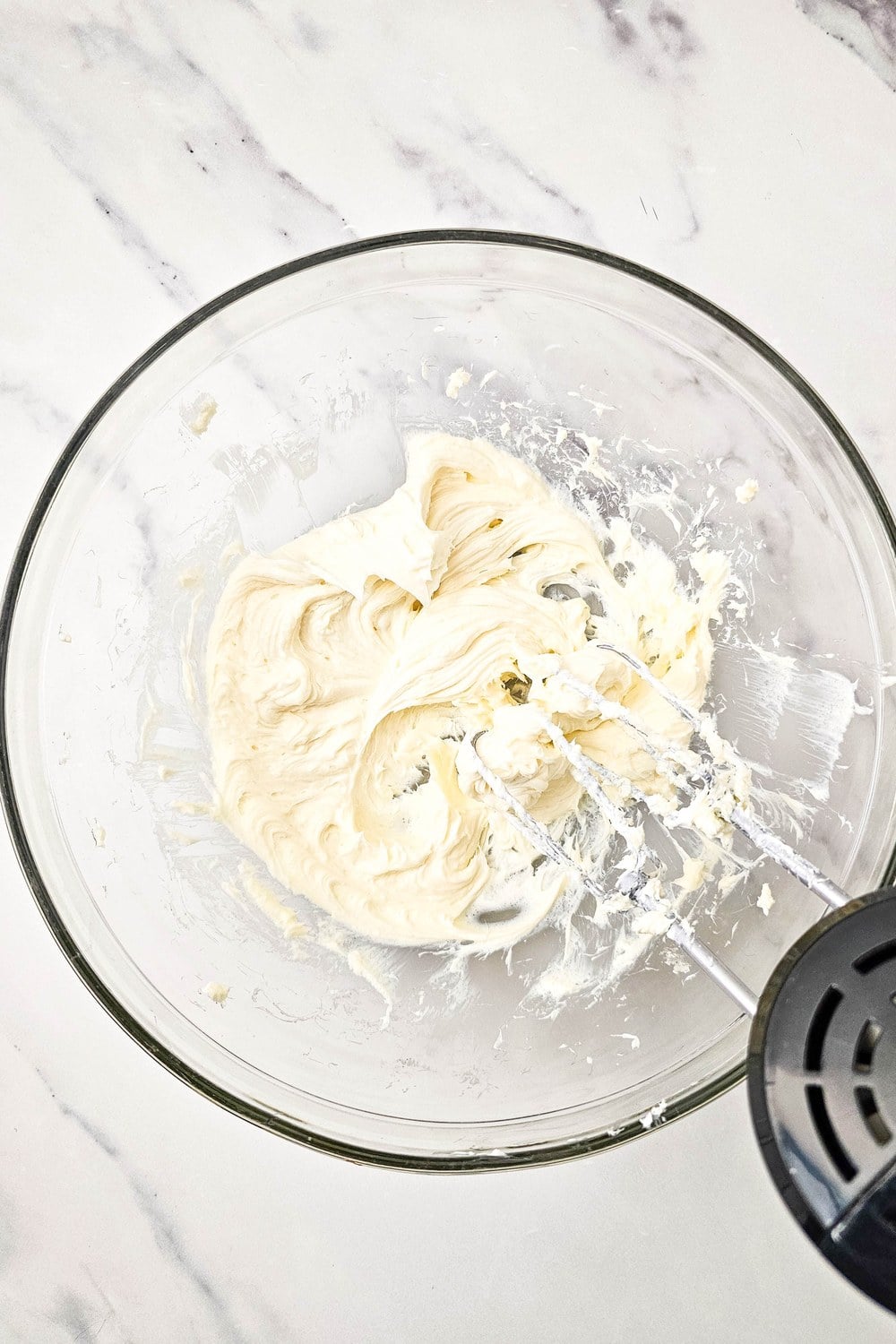 Smooth cream cheese frosting in a mixing bowl with hand mixer beaters, ready to spread on the sugar cookie crust