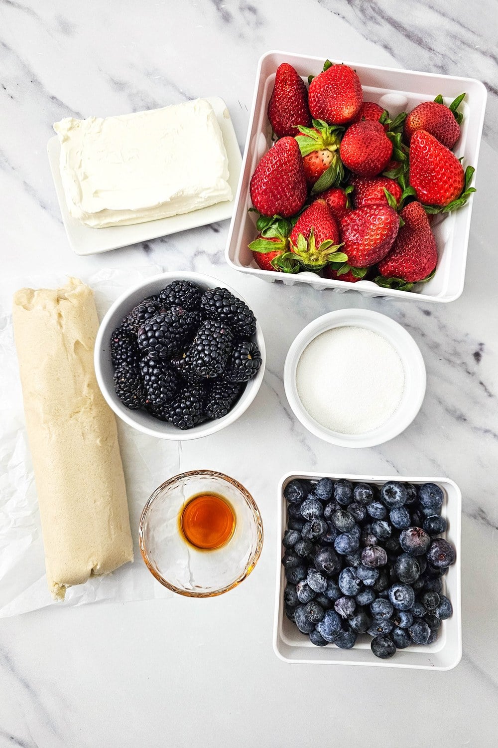 Flat lay of ingredients for red, white, and blue fruit pizza: cream cheese, sugar, vanilla, sugar cookie dough, strawberries, blueberries, and blackberries on a white marble surface