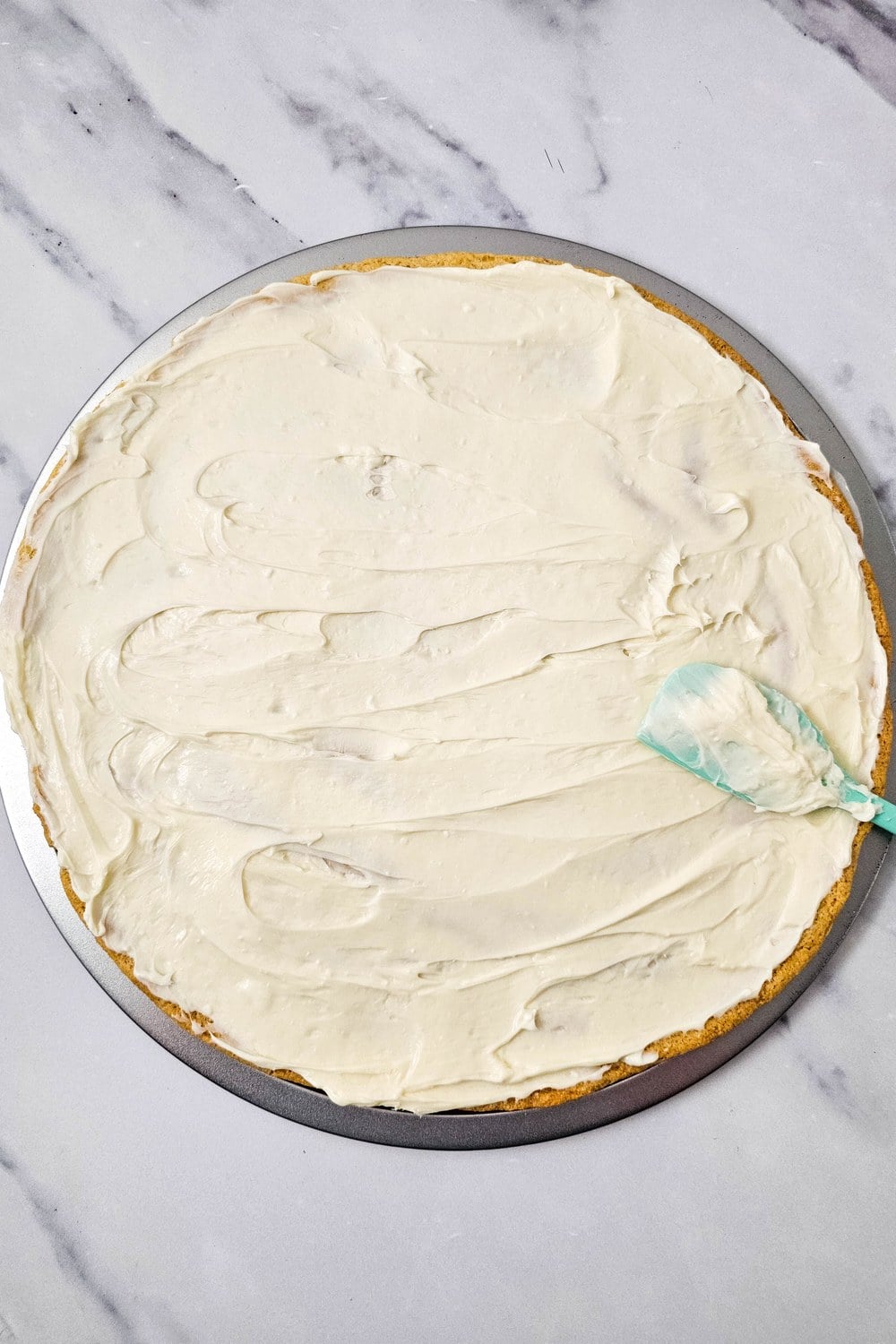 Cream cheese frosting spread edge to edge over a cooled sugar cookie crust with a teal silicone spatula
