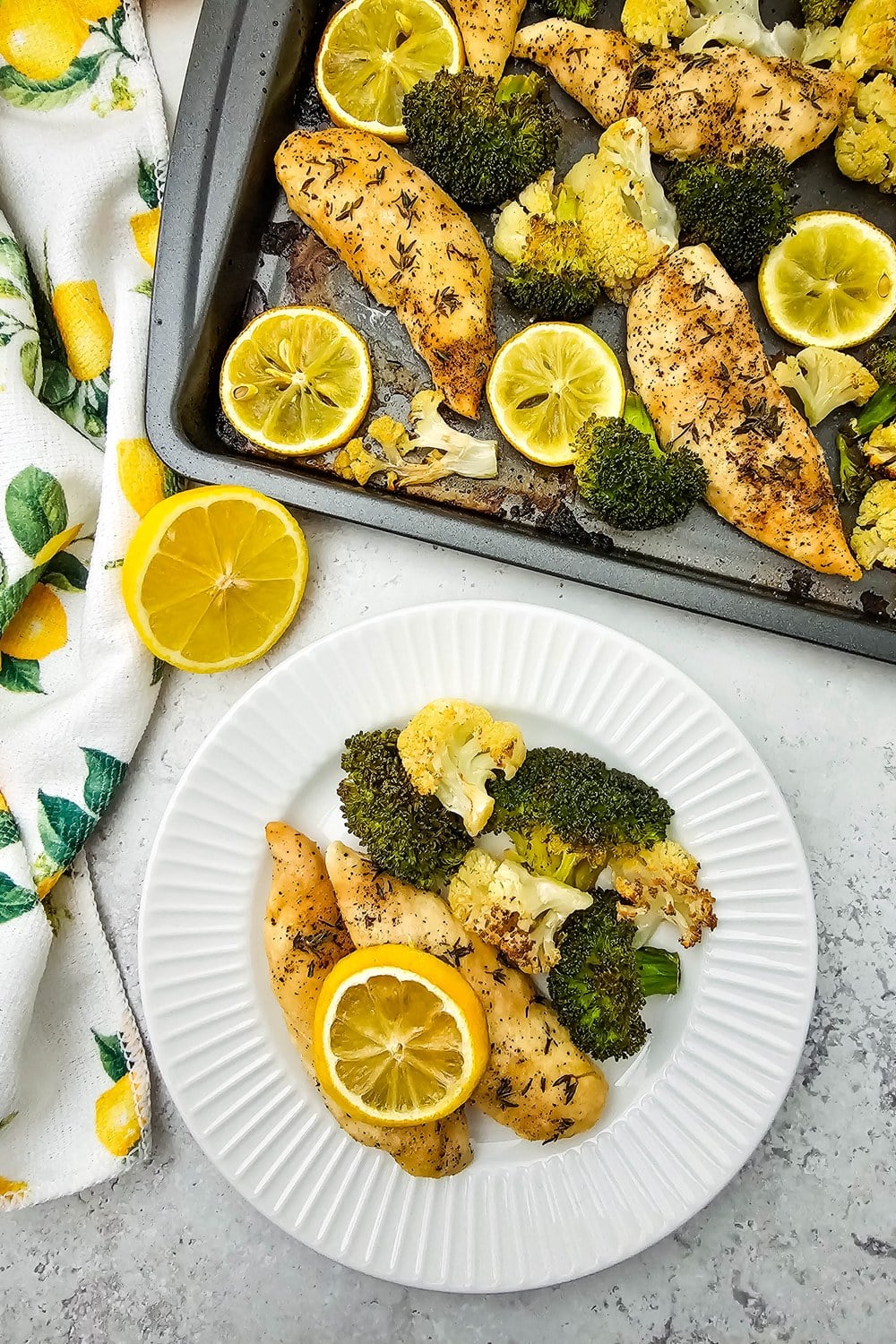 Plated sheet pan lemon chicken with a half-eaten sheet pan in the background and a lemon-print kitchen towel on the side.