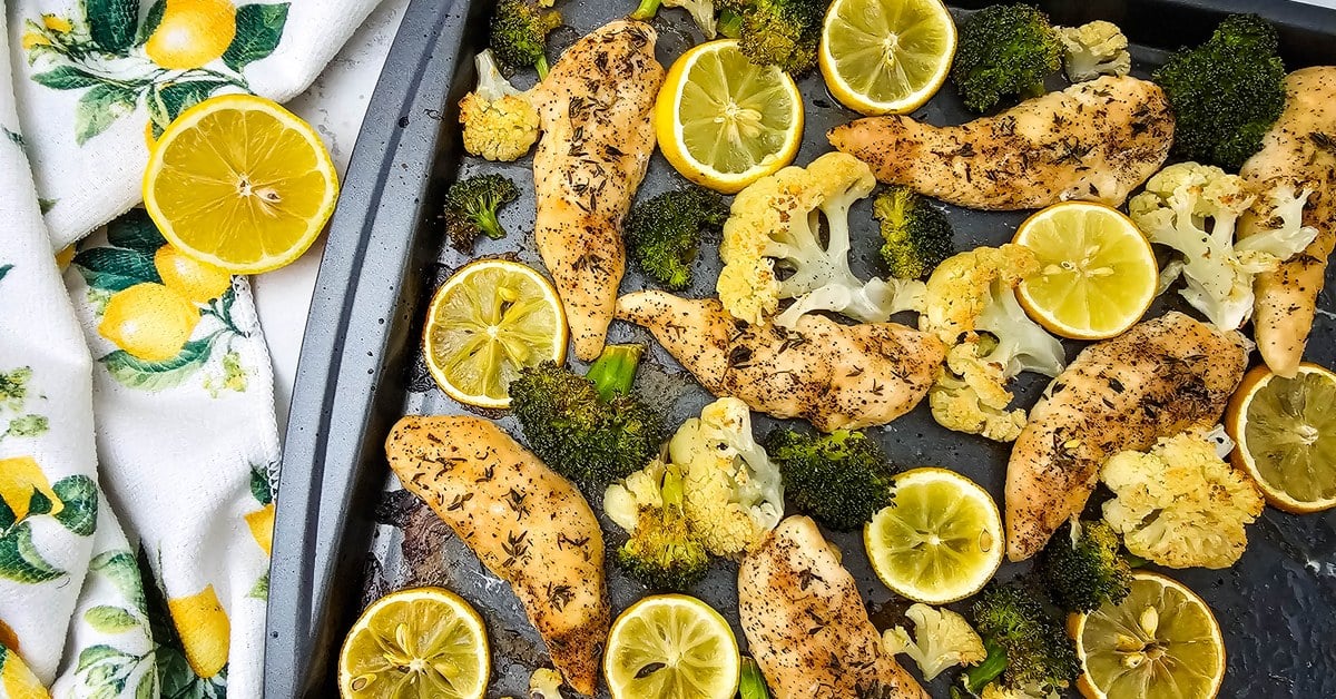 Wide overhead view of a sheet pan dinner with golden lemon chicken tenderloins, charred broccoli florets, roasted cauliflower, and bright lemon slices.
