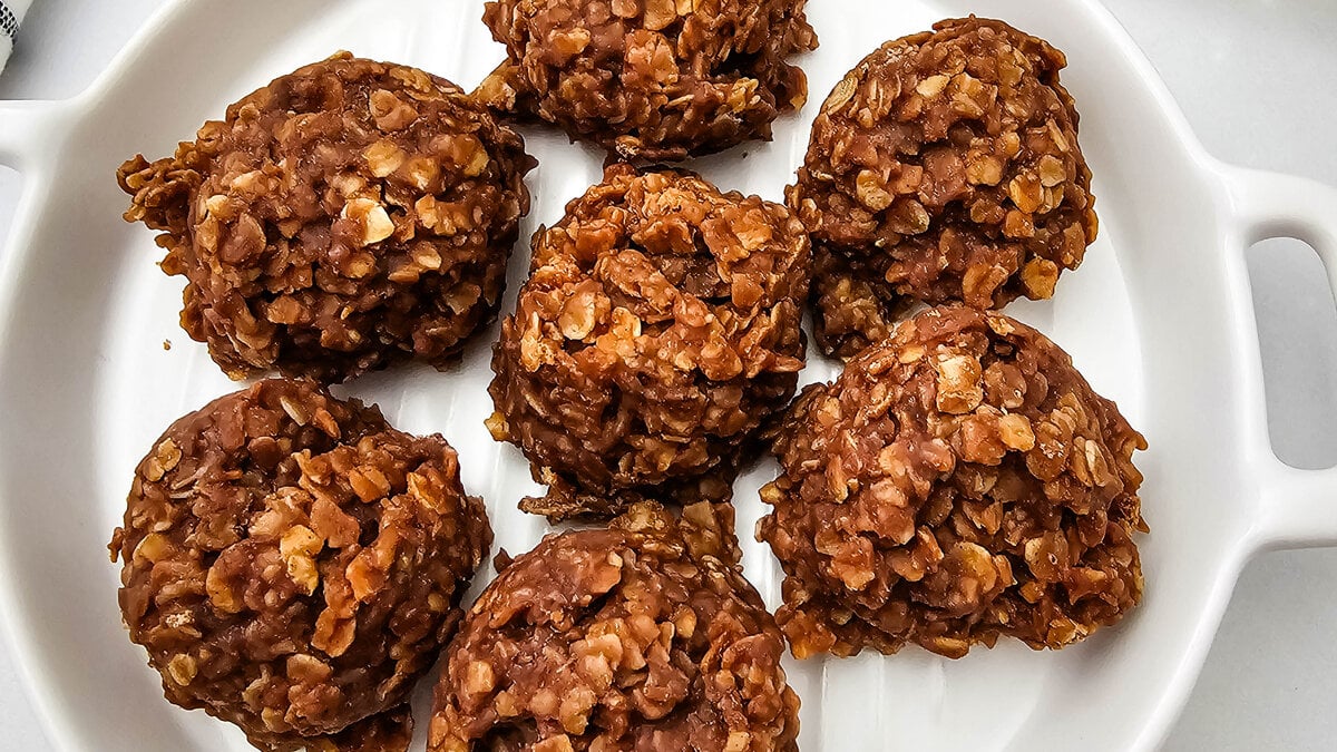 A plate of classic no-bake cookies with chocolate, peanut butter, and oats, more cooling on a rack behind.