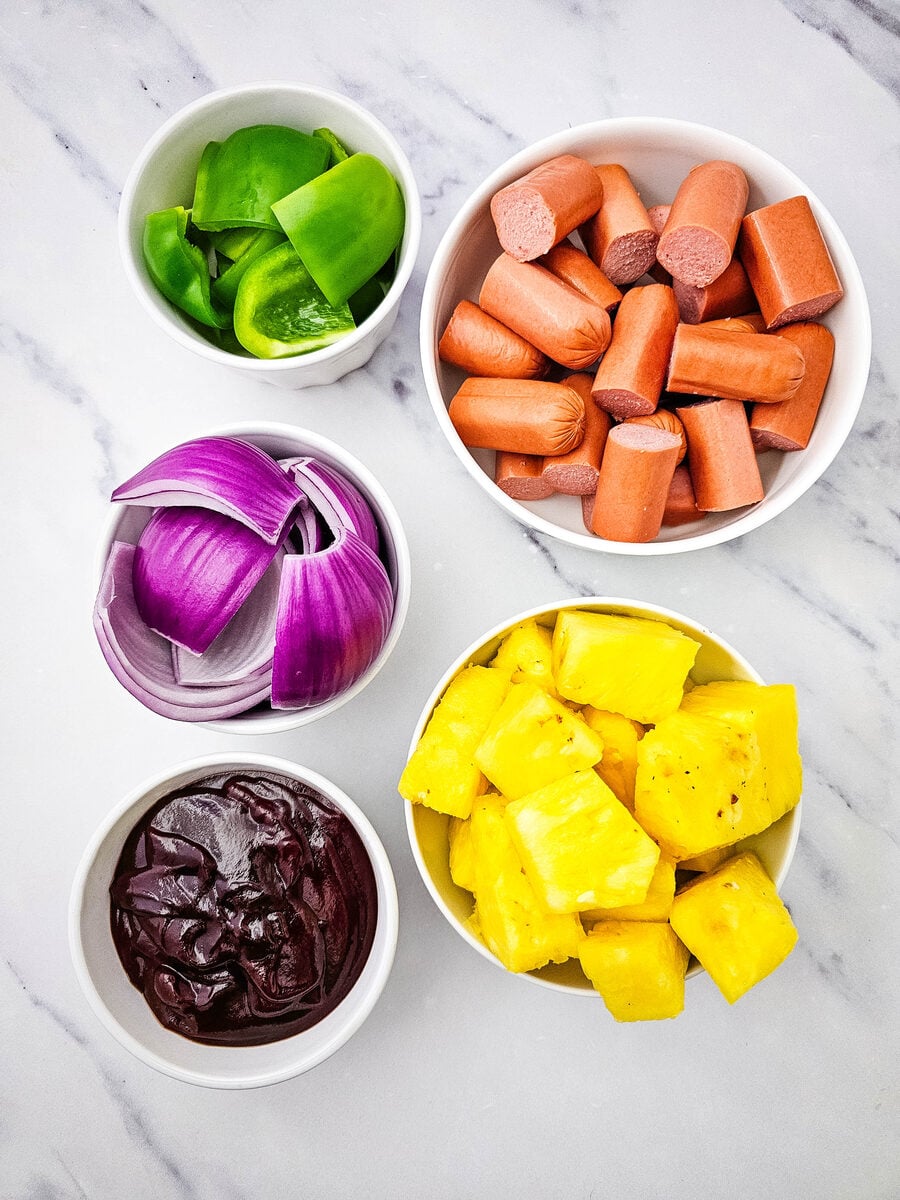 Ingredients for grilled hot dog kabobs including hot dogs, pineapple, green pepper, red onion, and barbecue sauce