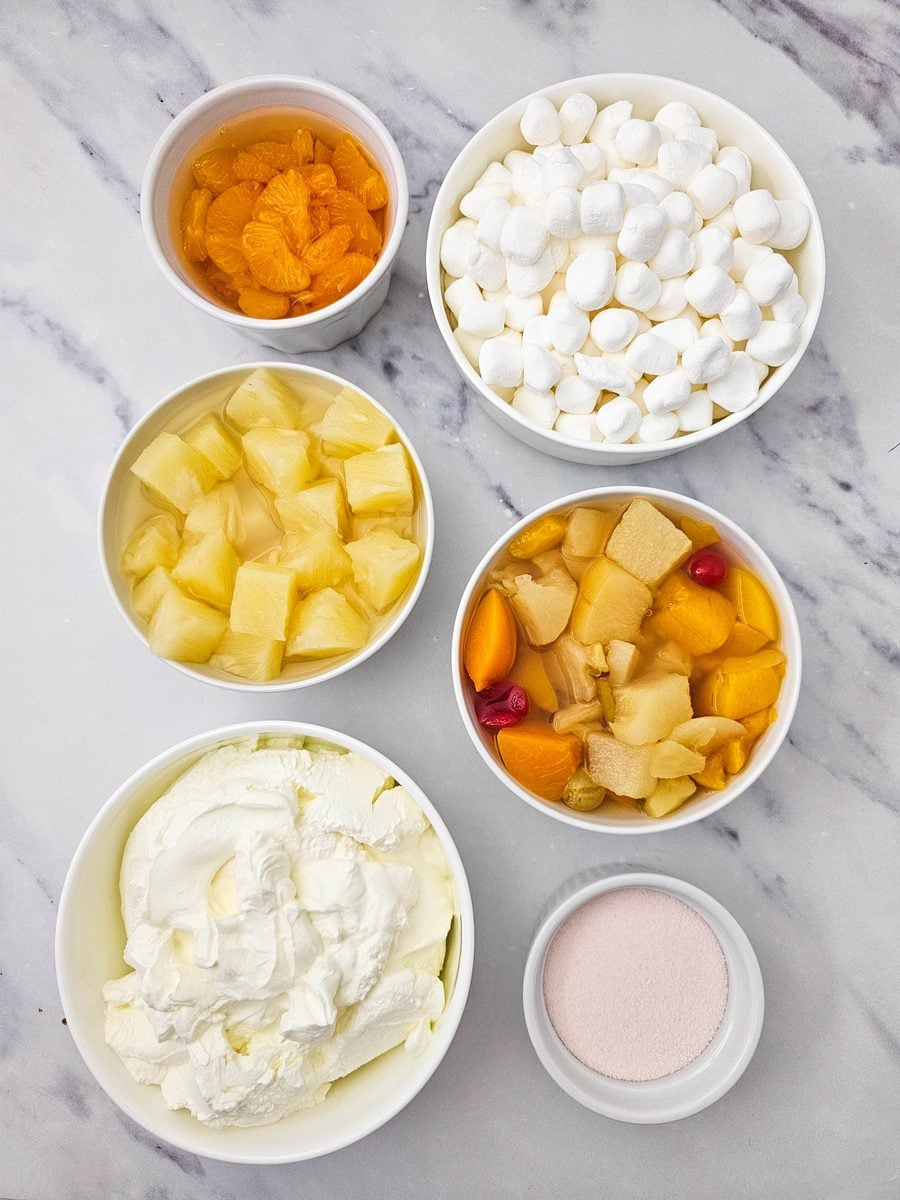 Hawaiian fruit salad ingredients in bowls: canned fruit, marshmallows, sour cream and orange gelatin