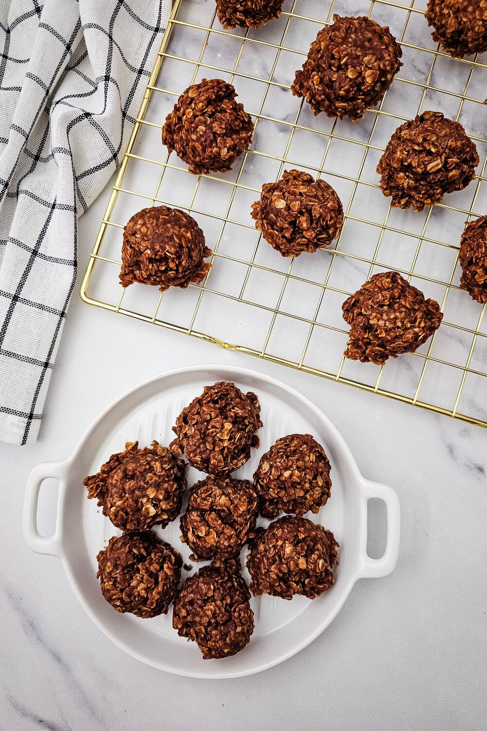 No-bake cookies setting on a gold cooling rack next to a plate of finished cookies.
