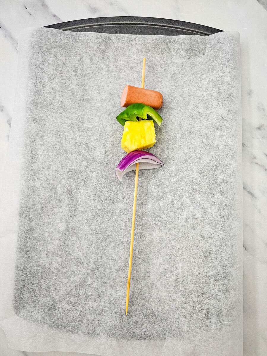 Threading hot dog, pineapple, pepper, and onion onto a skewer